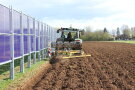 Das Foto zeigt einen Traktor beim pflügen der Ackerfläche neben einer vertikalen Agri-PV-Anlage.